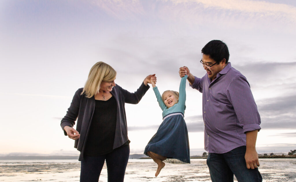 mom and dad swinging toddler daughter at Alameda crab cove beach.