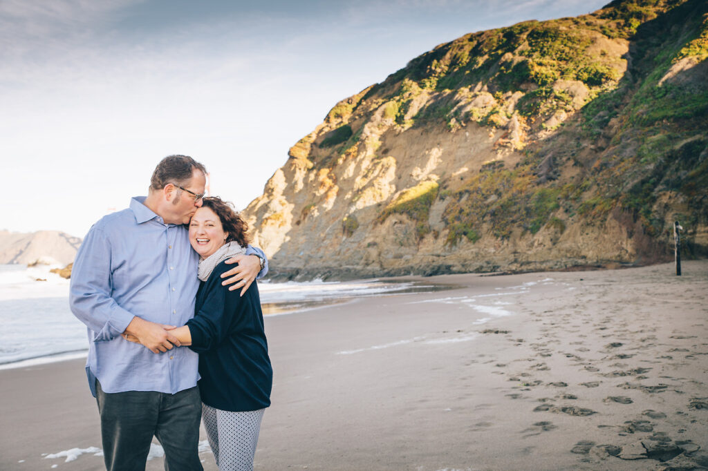 husband and wife hugging and laughing at baker beach during a family photo session.