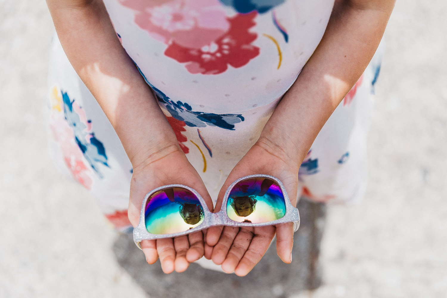 girl in colorful floral dress holding a pair of sunglasses with her reflection in them
