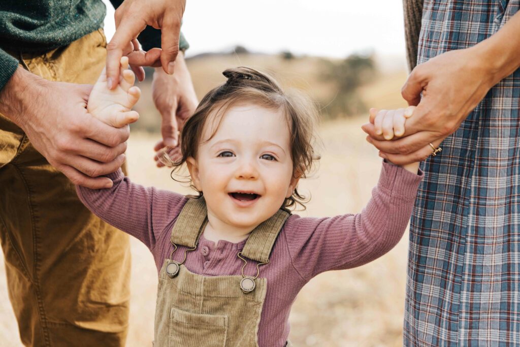 east-bay-family-photographer-IMG_9998-carmen-dunham-photography young toddler girl smiling while holding parents hand in meadow
