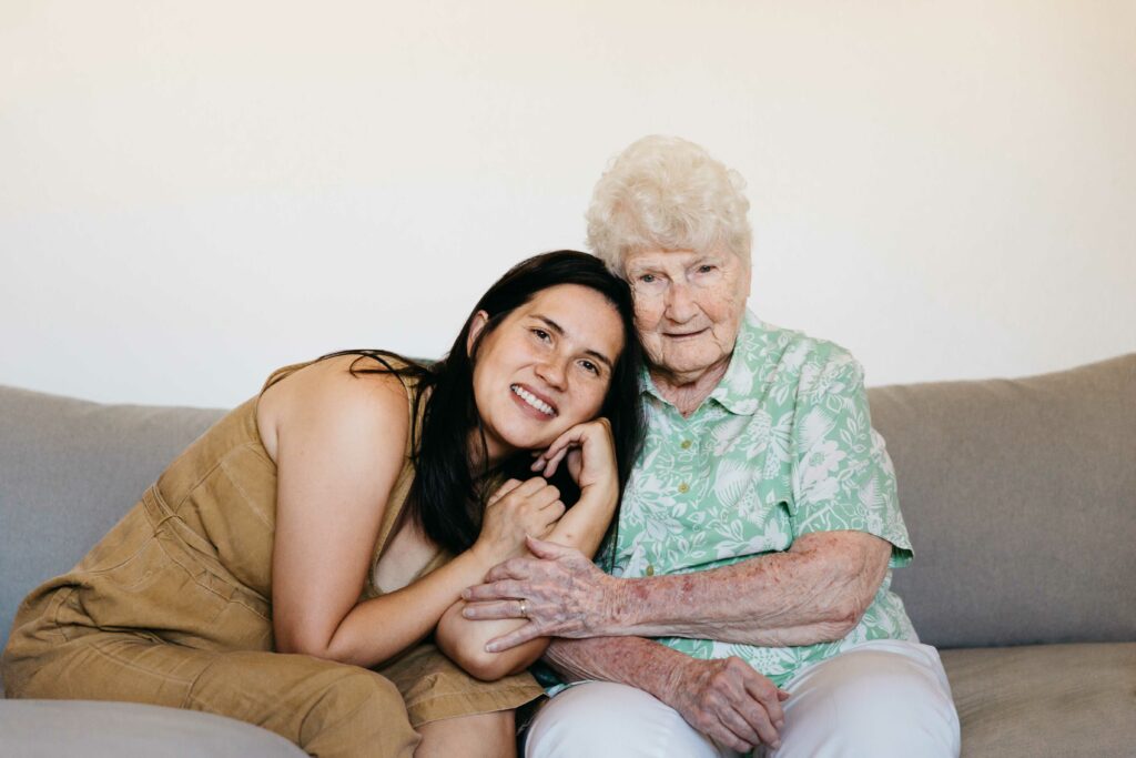 Adult granddaughter and grandmother holding each other on living room sofa