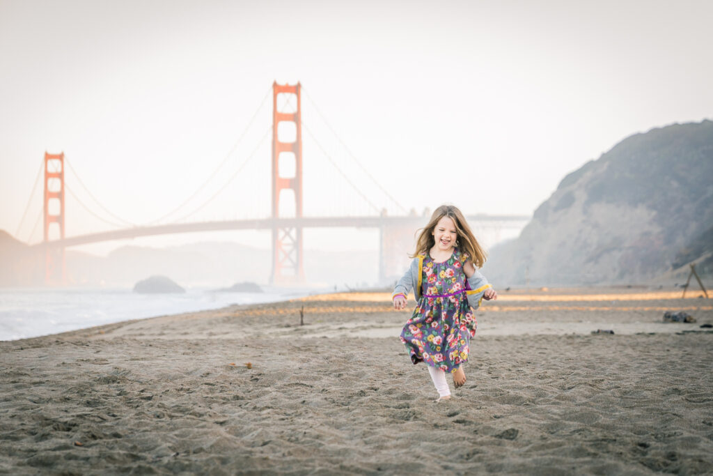 young girl running on baker beach with the golden gate bridge in the background.