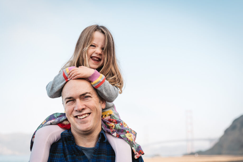 dad smiling while carrying young daughter on his shoulders during a family photo session at baker beach.