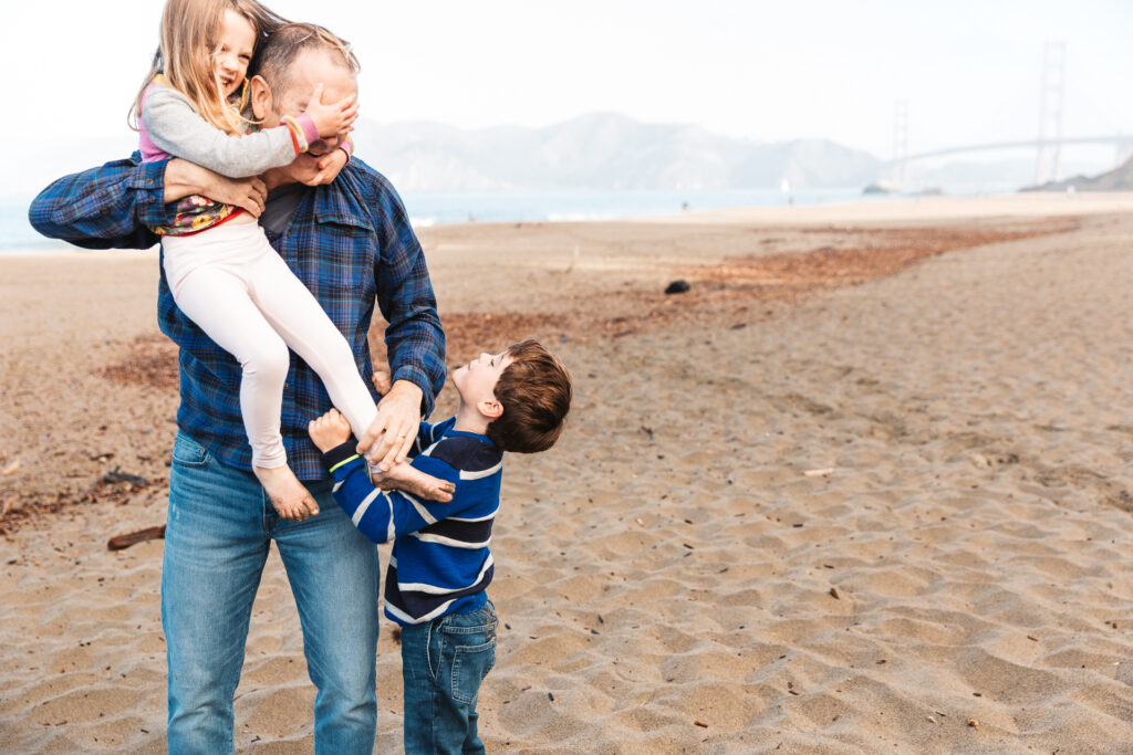 dad fumbling with young daughter and son during a family photo session at baker beach in san francisco.