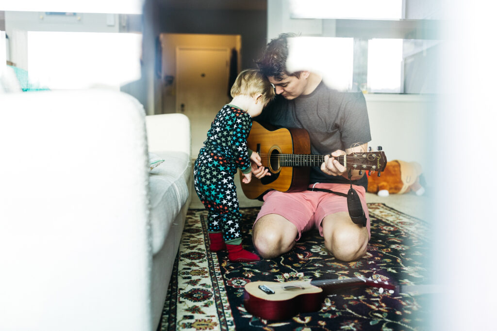 Dad playing the acoustic guitar to his toddler in the living room.