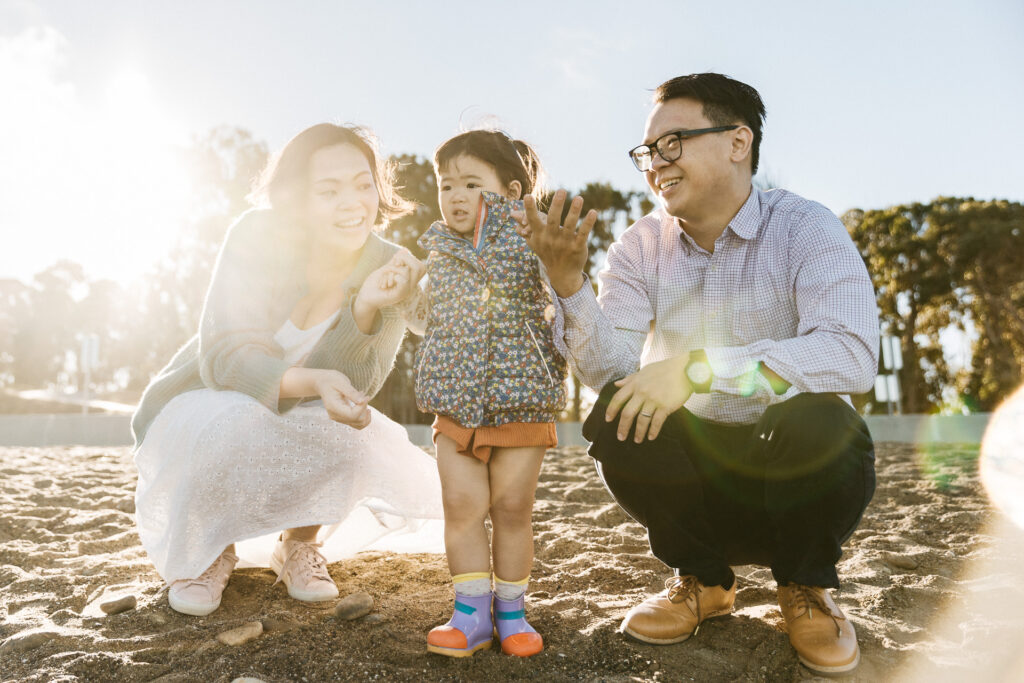 mother, toddler daughter, and husband holding hands on the beach as they look at the ocean.