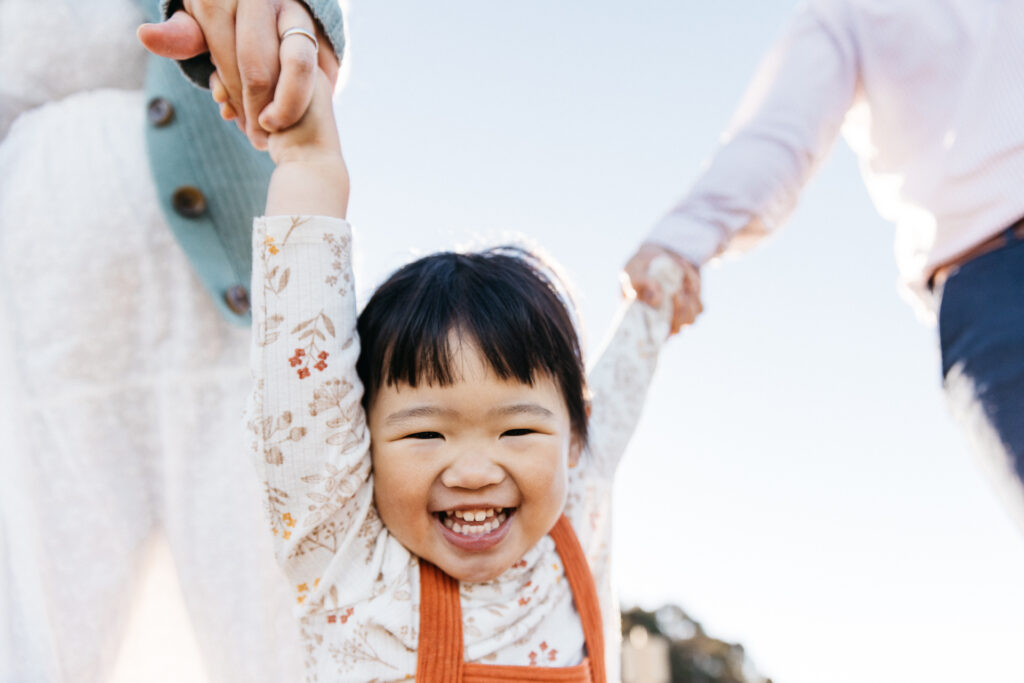 Toddler girl smiling big while holding mother and father's hands.