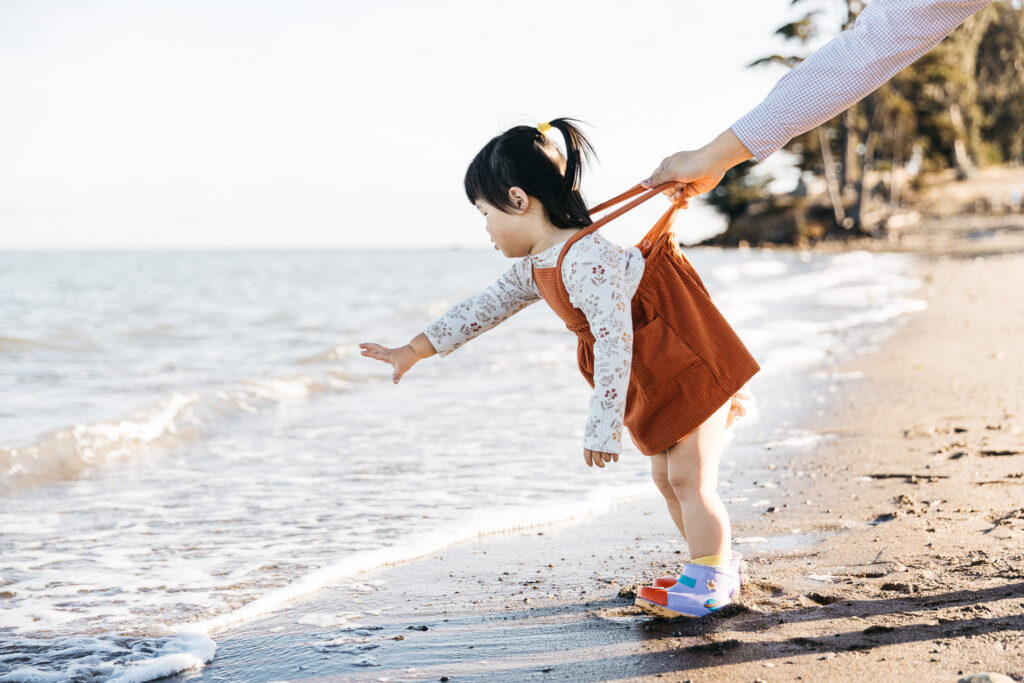 Young toddler girl reaching for the waves as her father holds her tightly by the straps of her dress.