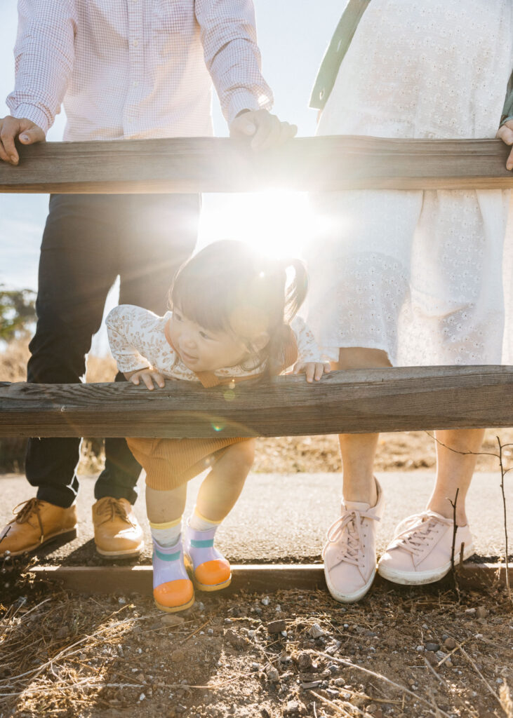 Toddler girl is hanging over wooden fence at the park.