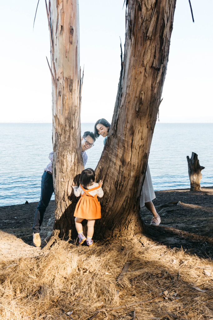 toddler girl is playing hide and go seek with her parents at a tree in Coyote Park.