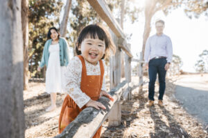 Young toddler girl is leaning against a wooden fence with her parents laughing in the background.