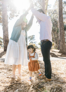Mom and dad are holding their hands in the air to form a tunnel for their toddler daughter to run through.