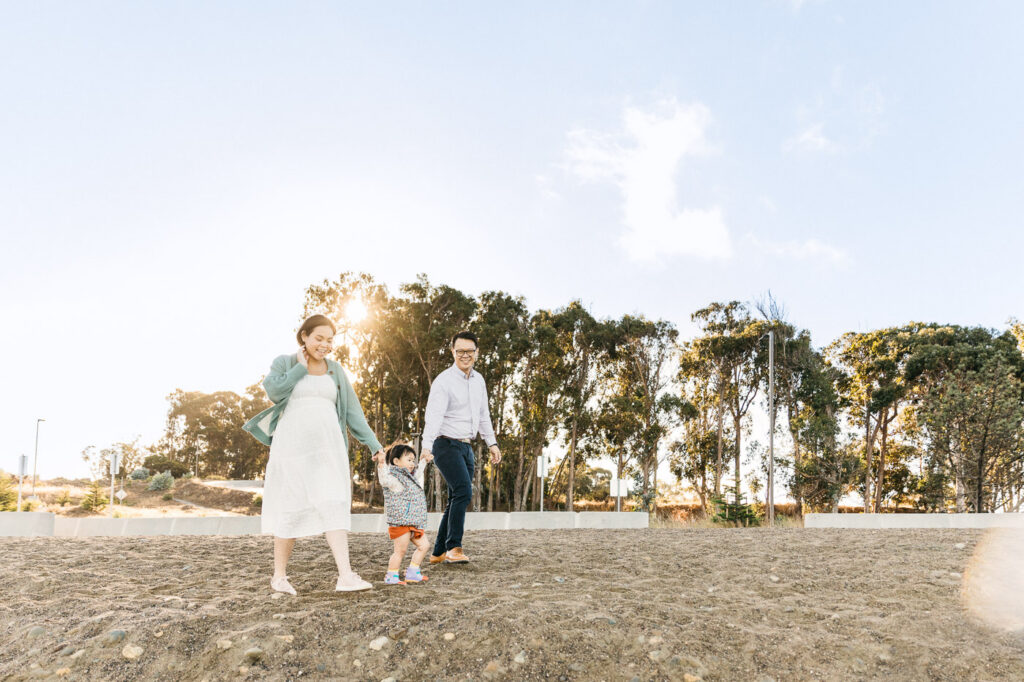 Mom and dad are walking on the beach while swinging their toddler daughter by the arms.