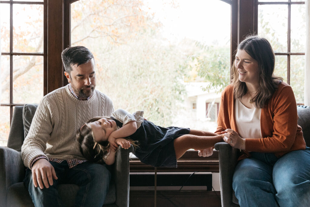 young girl balancing between two chairs while mom and dad look in amusement.
