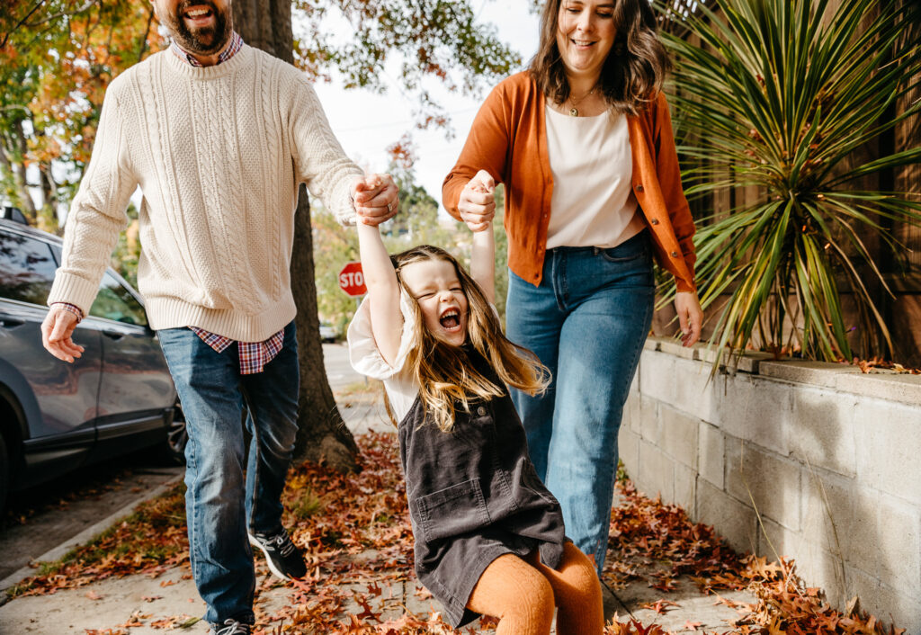 mom and dad swinging young daughter on the sidewalk.