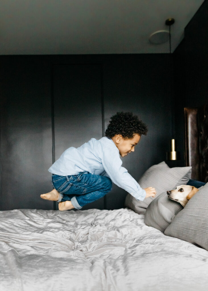 toddler boy jumping on the bed with his dog.