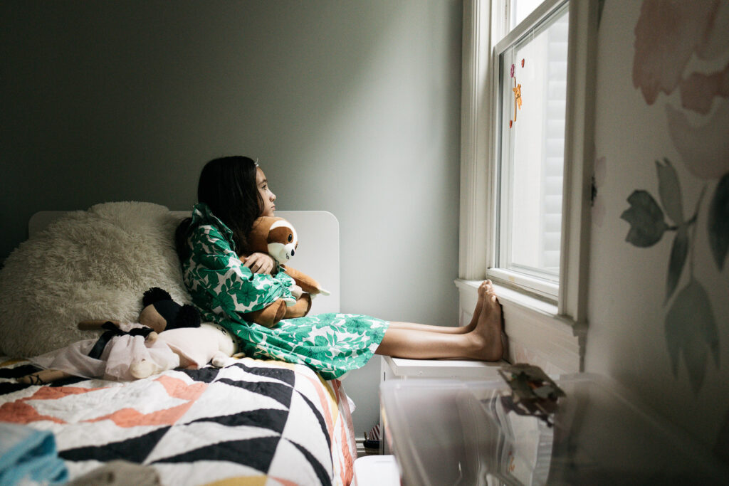 Girl is hugging her favorite stuffed animal sloth while looking out the window.