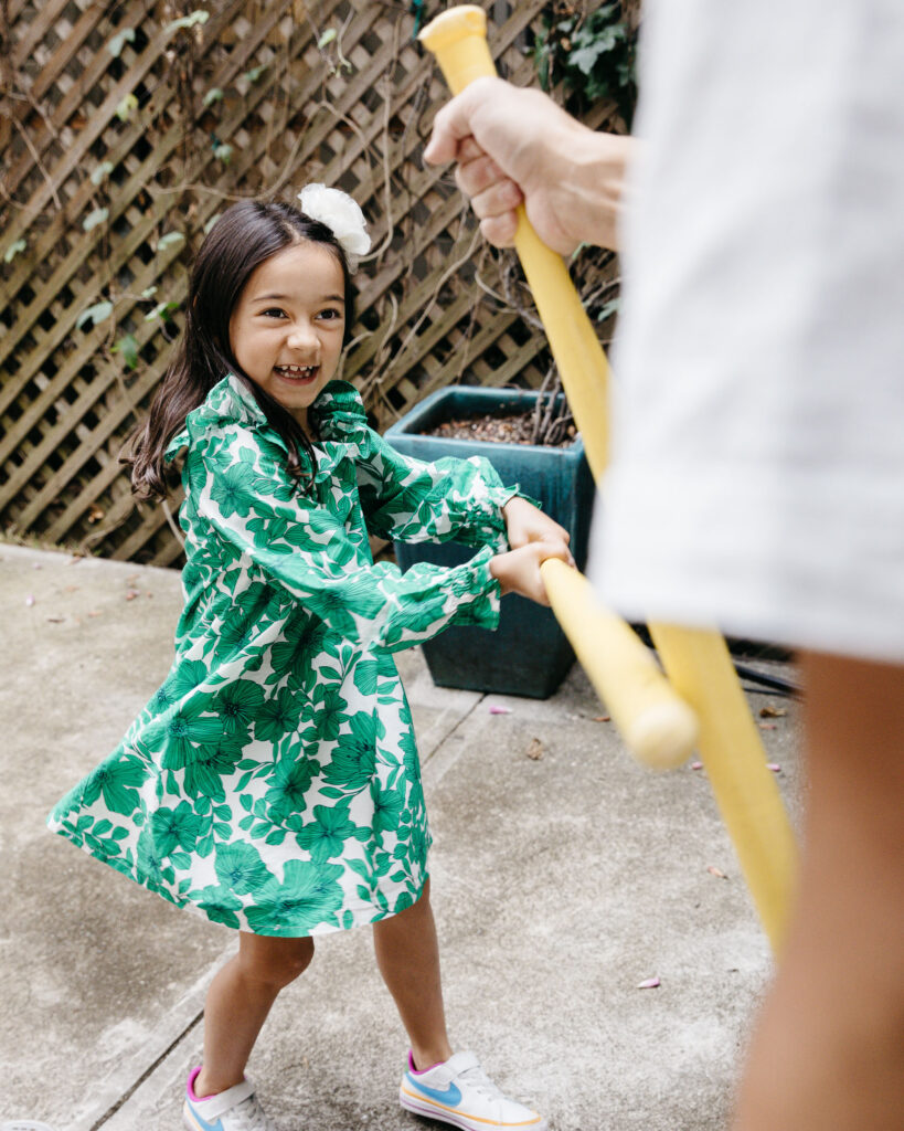 Eight year old is playfully hitting dad with a baseball bat.