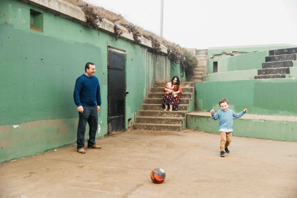 Dad and young toddler son are playing soccer at Battery Chamberlin while mom and newborn watch.
