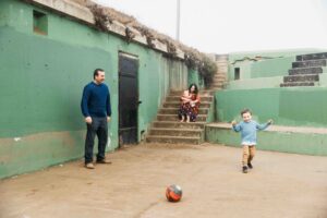 Dad and young toddler son are playing soccer at Battery Chamberlin while mom and newborn watch.
