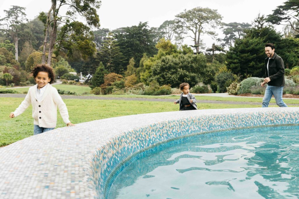 Dad is chasing his young toddler kids around the fountain during a family photo session.