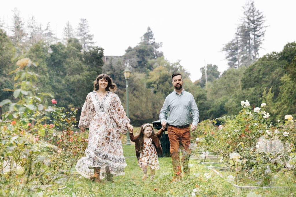Mom, dad, and young toddler daughter are walking and holding hands in the rose garden.