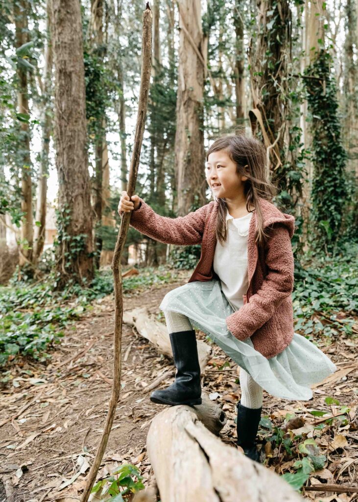 Young girl is confidently standing on a fallen tree branch holding a found tree branch as a staff.