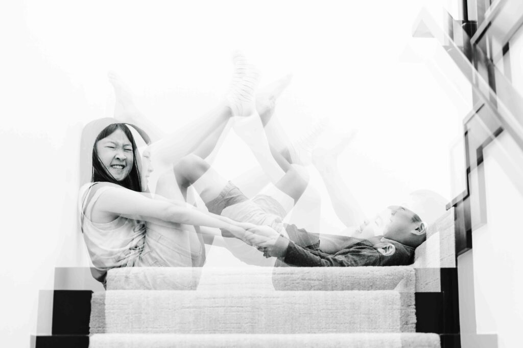 Young siblings are holding hands and flailing their legs at each other.