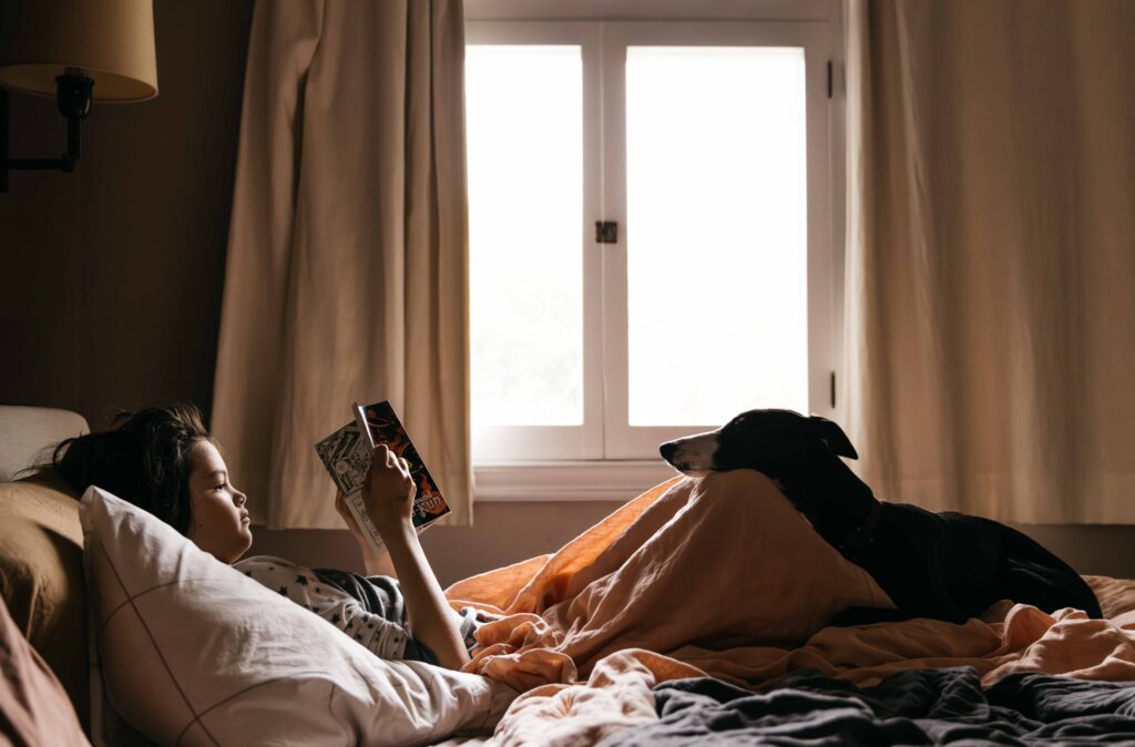 Teen girl is laying in bed reading a book with her whippet dog also resting its head on her knees.