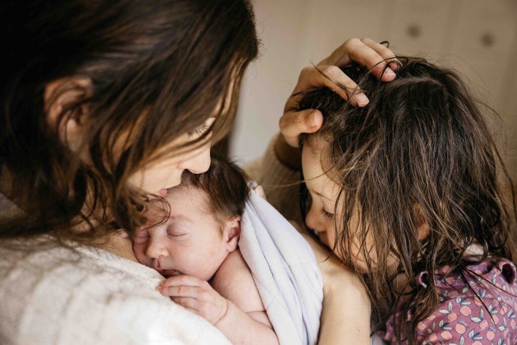 Mom is holding newborn baby on her chest while also running her fingers through the hair of her toddler girl.