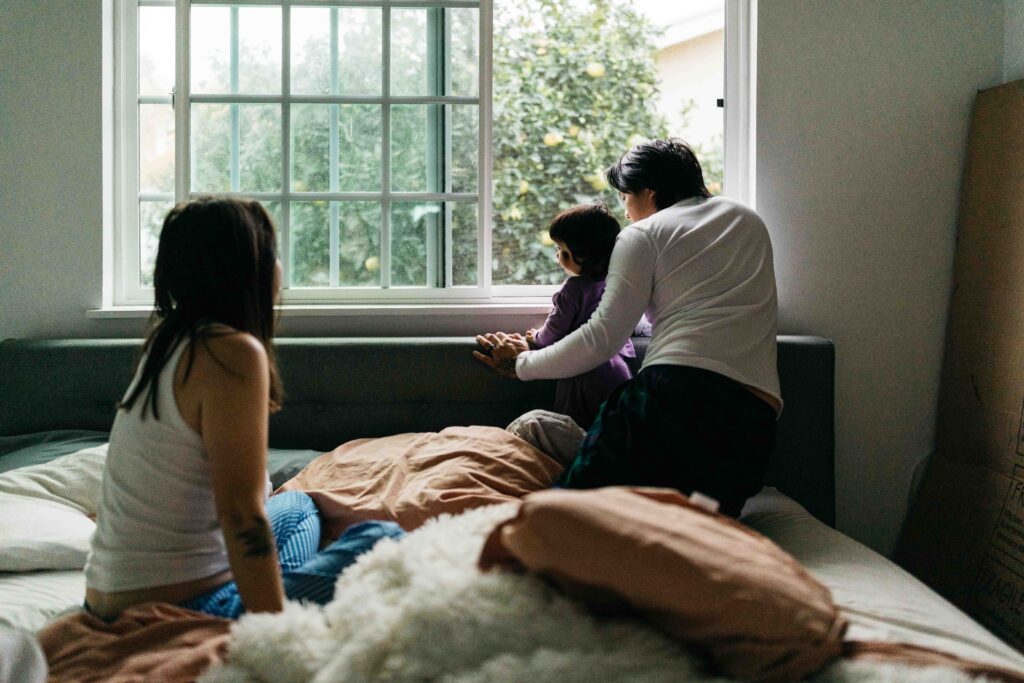 Mom is looking at husband and baby daughter as they gaze out the window in bed.