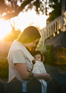 Mother is holding baby girl in a sling during a sunset newborn session at home in the front yard.