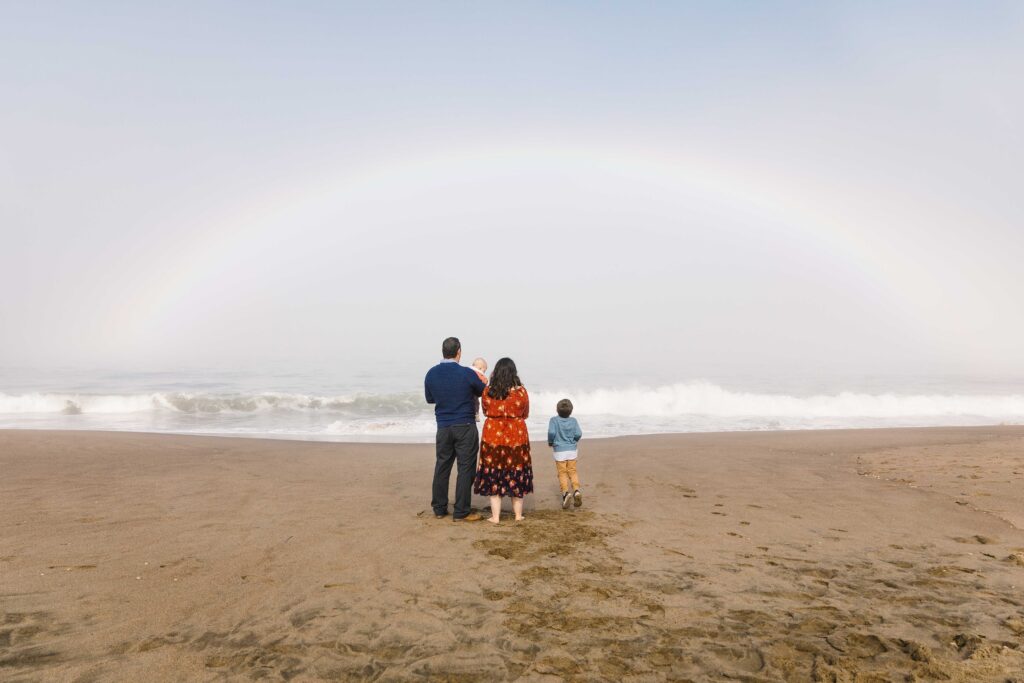 A family is looking out at the waves at the beach with a magnificent rainbow in the sky.