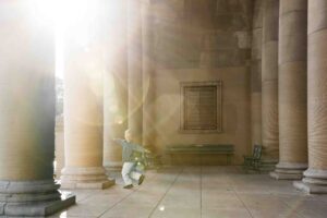 Young boy is dancing and weaving with his arms outstretched between the marble columns during a family photo session in Golden Gate Park.