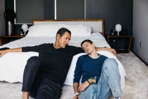 Dad and son are sitting at the foot of the bed relaxing.