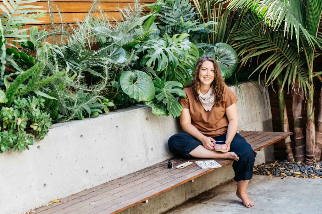 Ceramic artist sits outdoors with lush plants while drinking coffee and sketching.
