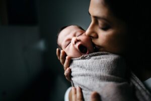 Mom is holding her swaddled newborn baby and kissing her gently on the cheeks.