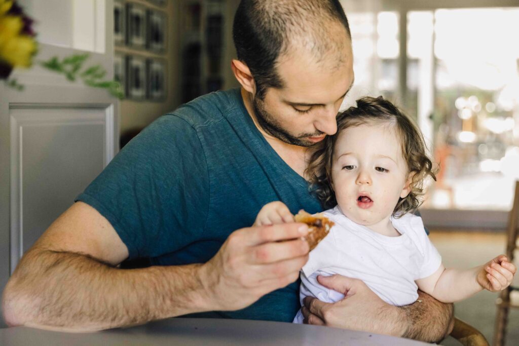 Dad holds toddler daughter on his lap while he feeds her during a newborn photo session.