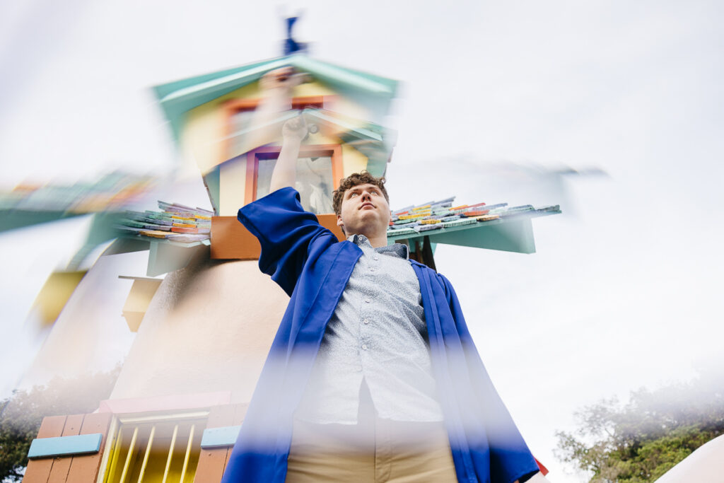 High school senior is waving his cap while wearing his graduate gown at the gates of Fairyland.