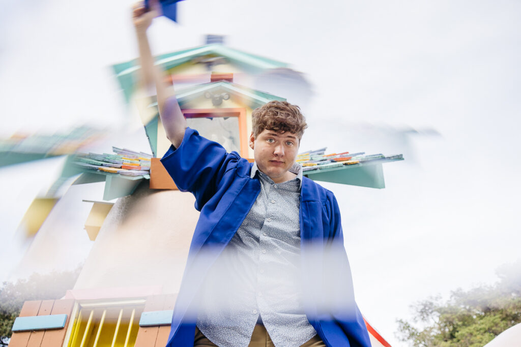 High school senior is waving his cap while wearing his graduate gown at the gates of Fairyland.