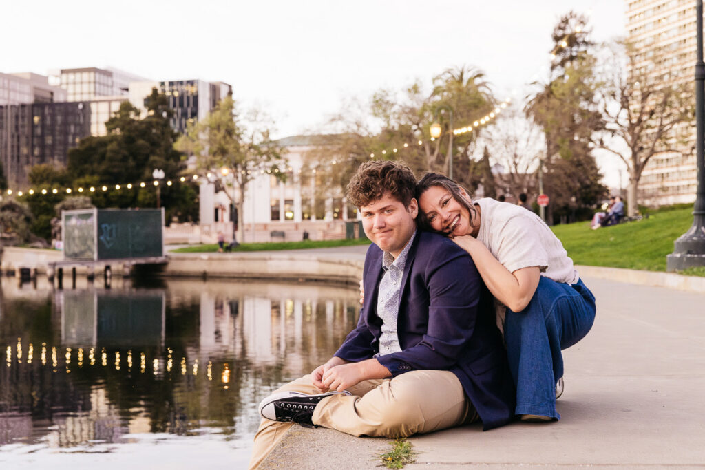 Young adult graduate is sitting at Lake Merritt while his mom leans gently against his back during a family photo session.