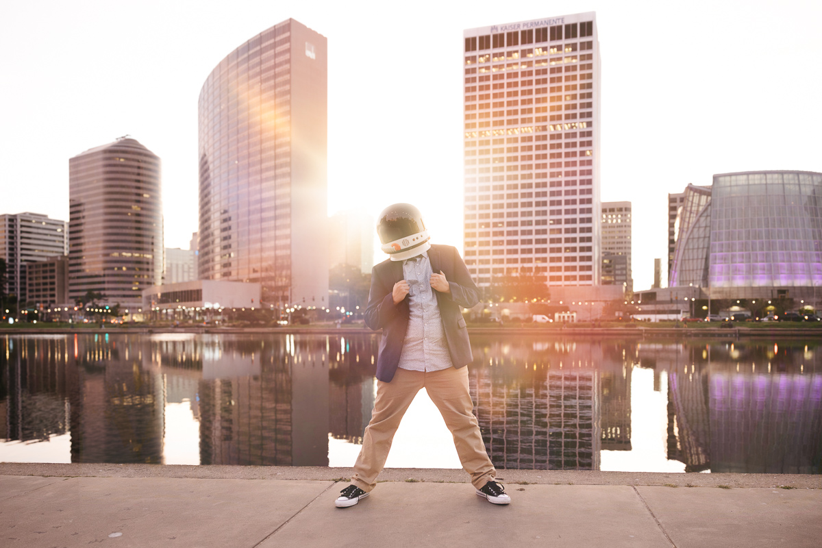 Senior graduate is standing at Lake Merritt during a spectacular sunset wearing his suit and astronaut helmet.