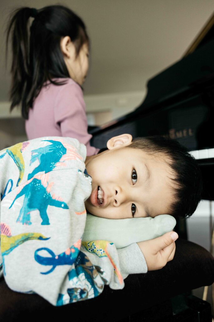 Toddler brother is laying on the piano bench while his sister plays at the piano.