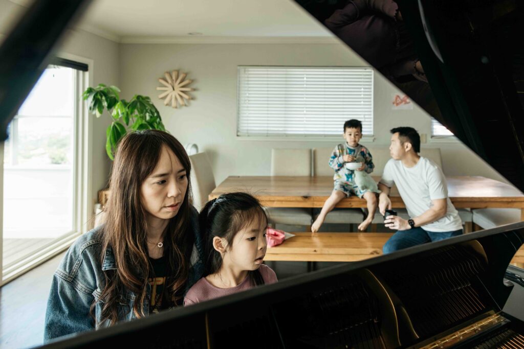 Mom and daughter are playing at the piano while husband and brother watches.