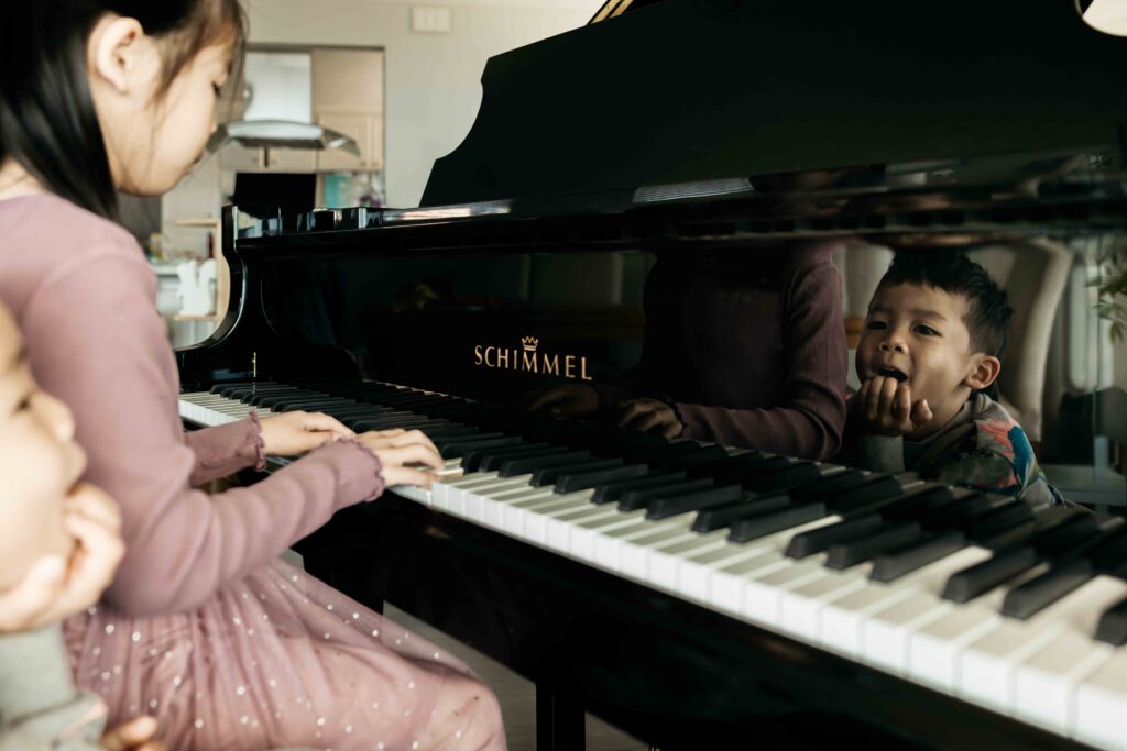 Younger brother is looking at his reflection in the piano while his sister plays the piano.