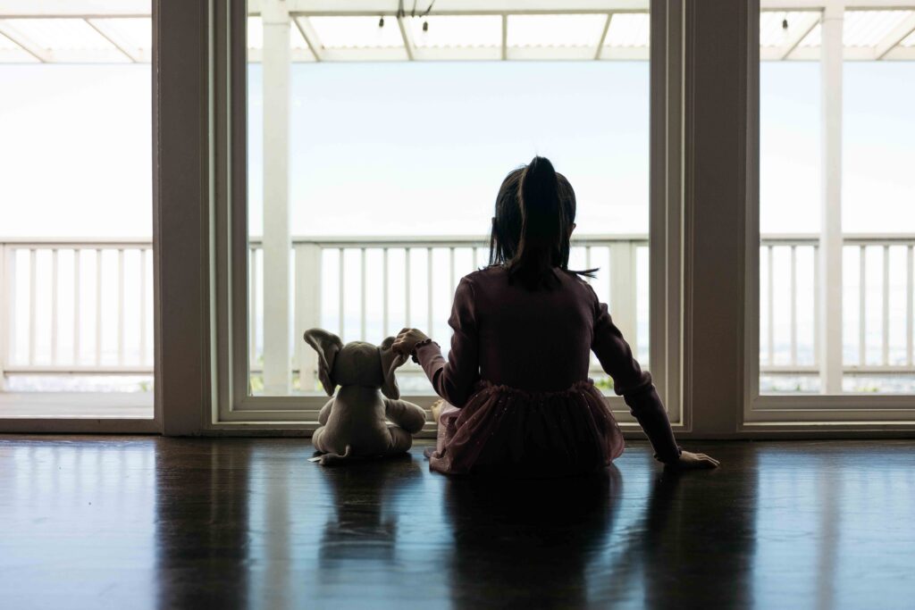 Toddler girl is sitting next to her elephant stuffed animal at the window.