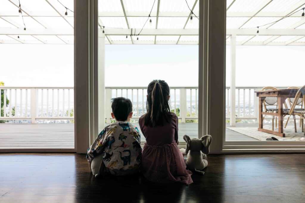 Sibling brother and sister is sitting at the window with a stuffed elephant.