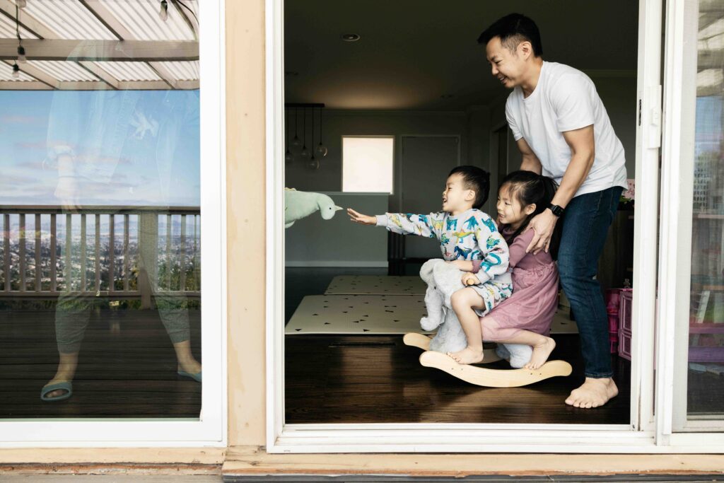 Toddler siblings are sitting on a rocking horse with dad pushing them during a family photo session at home.