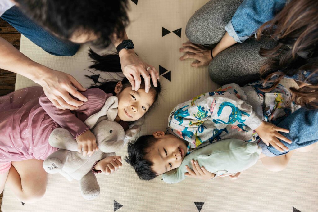 Siblings are laying on the ground holding their favorite stuffed animals while their parents hold them.