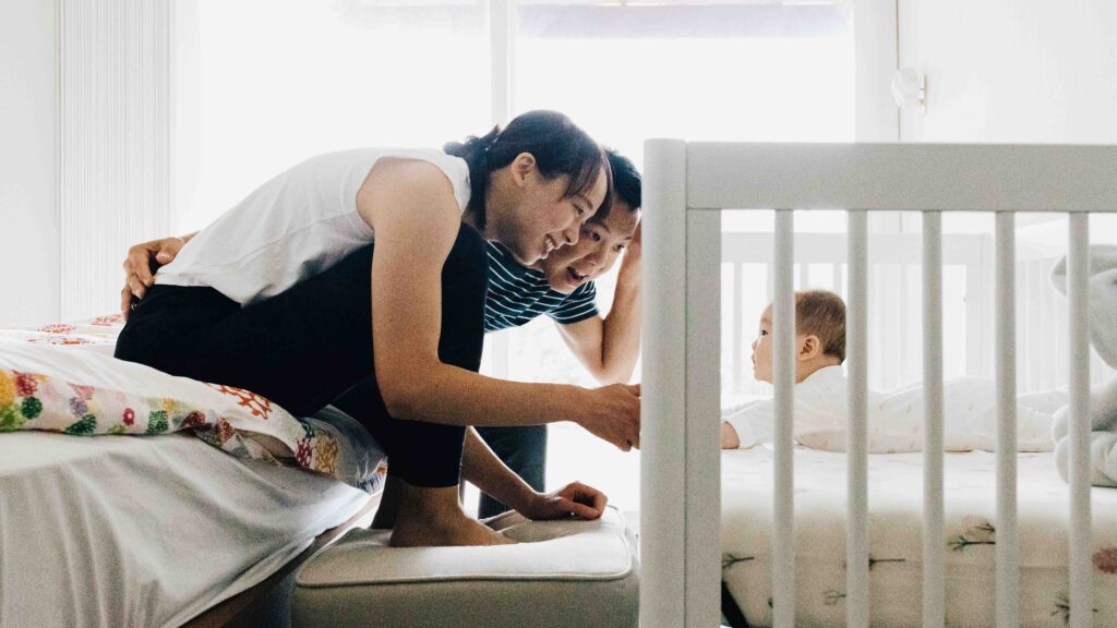 Mom and dad are playing peek-a-boo with baby through the crib slats during a newborn photo session at home.